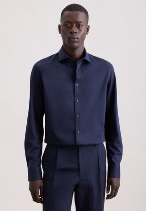 Homme portant une chemise cintrée à manches longues bleu foncé avec des boutons et un pantalon assorti, debout devant un arrière-plan clair uni.