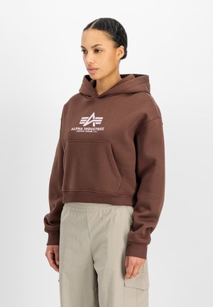 Junge Frau trägt einen braunen Alpha Industries Hoodie und hellbeige Cargohosen, steht vor einem schlichten weißen Hintergrund.