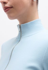 Ανοιχτό μπλε ριγωτό πουλόβερ με φερμουάρ και ψηλό γιακά, με ασημί λεπτομέρεια φερμουάρ. Κοντινή λήψη που εστιάζει στη  γραμμή του λαιμού και την υφή του υφάσματος.