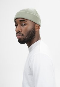 Gehaakte beanie in zacht groen, met ribbelstructuur en een subtiele naadgetailleerde detail op de kroon. Gedragen met een aansluitend wit langemouwen shirt.