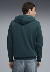 Sweat à capuche vert foncé au design ajusté, avec une capuche, des poignets côtelés et un tissu texturé. Porté avec un jeans en denim bleu clair.