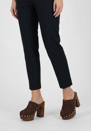 Persoon die zwarte slim-fit broek draagt en bruine platformklompen met luipaardprint, houten dikke hakken en gouden studs.
