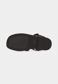 Semelle de sandale noire avec une zone de grip texturée près des orteils, talon lisse, et une boucle de bride de cheville visible.