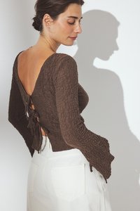 Top marrone a trama con scollatura a V profonda, dettaglio con lacci e maniche lunghe. Abbinato a pantaloni bianchi a vita alta. Ombria visibile sullo sfondo.