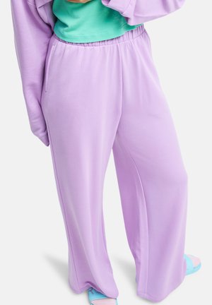 Persoon draagt loszittende lavendelkleurige broek met zijzakken, gecombineerd met een groen top en pastelkleurige blauwe en roze sandalen op een witte achtergrond.