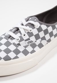 Låg sneakers med grå och vit rutmönster, vita snörningar, vit gummisula och Vans-logotagg på sidan.