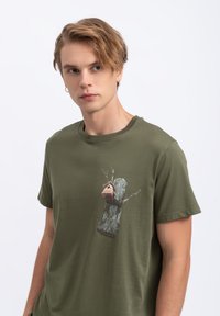 Olivengroene katoenen T-shirt met een grafiek van een boomstronk waaraan een vogelhuisje is bevestigd, gedetailleerd met takken en een gestructureerde schors.