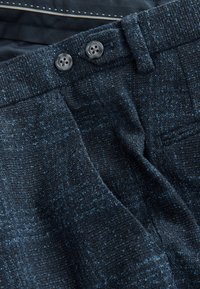 Tela texturizada azul oscuro con un patrón cuadriculado, con dos botones negros y una cintura lisa, parcialmente doblada.