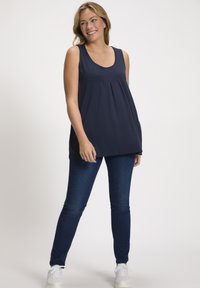 Top senza maniche blu navy con scollatura a barchetta, pieghe sul fronte e vestibilità rilassata, abbinato a jeans scuri in denim e sneakers bianche.