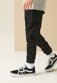 Pantaloni de trening negri cu buzunar lateral, manșete striate și textură moale, asortați cu adidași negri Vans și șosete albe până la gleznă.