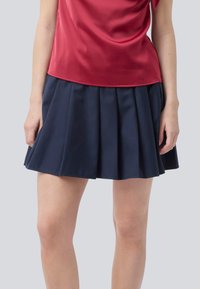 Femme portant une jupe plissée bleu marine et un haut sans manches en satin rouge, se tenant devant un fond gris clair uni.