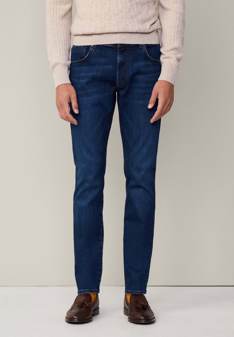Mænds mørkeblå jeans lavet af denim; slim fit med fem lommer, subtil syning, båret med en lysfarvet striktrøje og brune loafers.