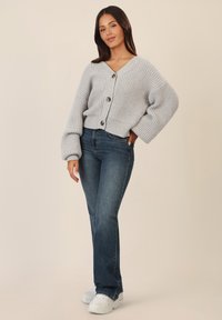 Cardigan in grigio chiaro a coste con scollatura a V e bottoni, abbinato a jeans a zampa d'elefante blu scuro e sneakers bianche su uno sfondo neutro.