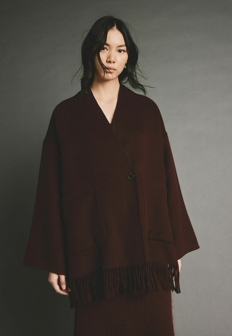 Burgundy színű, oversize kabát gyapjú keverékből, bojtolt aljjal, mély zsebekkel és egy gombos záródással.
