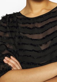 Svart blus med genomskinligt tyg med horisontella, strukturerade ränder och fransdetaljer. Mjuk, lager-på-lager design ger visuell intresse.