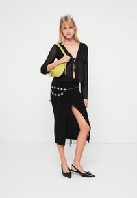 Top trasparente nero a maniche lunghe con dettaglio a fiocco, gonna nera con spacco laterale e catena argentata, abbinati a una borsa verde lime e scarpe nere.