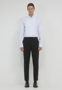 Uomo in piedi contro uno sfondo bianco, indossa una camicia da smoking bianca con pettorina, pantaloni neri su misura e scarpe nere eleganti.