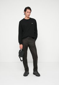 Μαύρο πουκάμισο με μακριά μανίκια και πράσινο λογότυπο, καστανές παντελόνες με σχέδιο, μαύρη τσάντα με λαβές και μαύρα αθλητικά παπούτσια. Το μοντέλο στέκεται μπροστά σε λευκό φόντο.