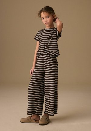 Young child standing, wearing matching black and white striped short-sleeve top and wide-leg pants, with tan slip-on shoes on neutral background.