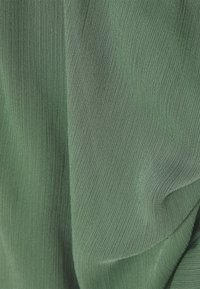 Tissu texturé vert avec de fines ondulations, suggérant un matériau léger. La couleur est une teinte de vert douce et atténuée.