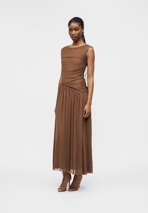 Kvinde med flettet hår iført en brun ærmeløs kjole med rynker og brune åbne tå-sandaler, stående mod en ensartet lys grå baggrund.