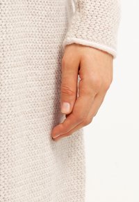 Gros plan sur une main aux ongles naturels reposant contre une manche en tricot de couleur crème.