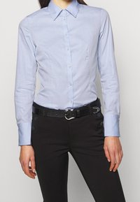 Camicia azzurro chiaro con colletto, realizzata in tessuto liscio, caratterizzata da un design aderente e chiusura con bottoni. Indossata con pantaloni neri e una cintura strutturata.