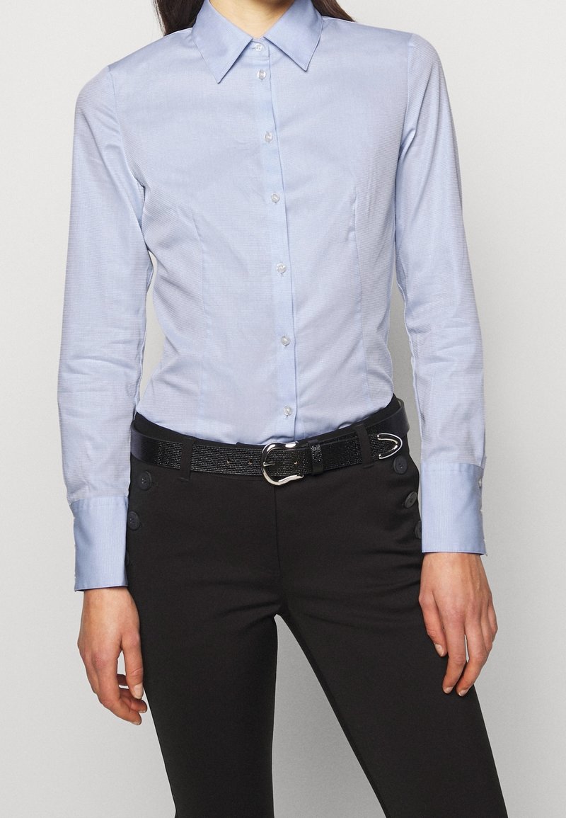 Camicia azzurro chiaro con colletto, realizzata in tessuto liscio, caratterizzata da un design aderente e chiusura con bottoni. Indossata con pantaloni neri e una cintura strutturata.