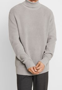 Mann trägt einen hellgrauen, gerippten Rollkragenpullover und dunkle grau Hosen, mit gefalteten Händen vor sich.