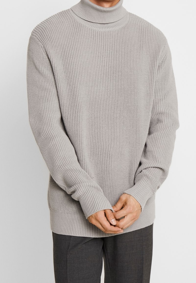 Mann trägt einen hellgrauen, gerippten Rollkragenpullover und dunkle grau Hosen, mit gefalteten Händen vor sich.