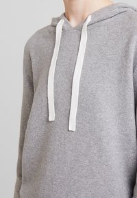 Sweat à capuche gris en tissu doux avec une coupe standard, doté d'un cordon de capuche blanc. La texture semble lisse et raffinée.