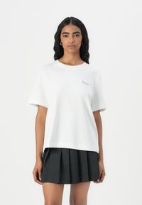T-shirt bianco con vestibilità rilassata, maniche corte e un piccolo logo nero "SELECTED" sul petto, abbinato a una gonna nera plissettata.