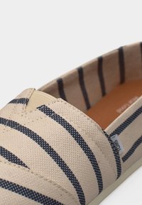 Μπεζ καμβαδένιο παπούτσι χωρίς κορδόνια με μαύρες οριζόντιες ρίγες και λαστιχένια σόλα, με μια μικρή υφασμάτινη ετικέτα μάρκας στο πλάι.