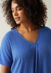 Blå tekstureret V-hals top med korte ærmer, der har et subtilt, krøllet mønster og blødt stof, der let draperer.