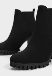 Black ankle boots made of soft suede, featuring a chunky rubber outsole with a jagged edge and a tapered high heel for stability.