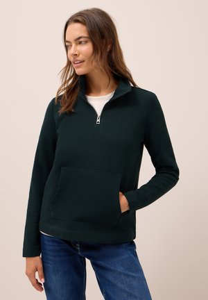 Femme portant un pull à zip quart vert foncé avec poche avant et jeans bleus, se tenant avec une main dans la poche, regardant sur le côté.