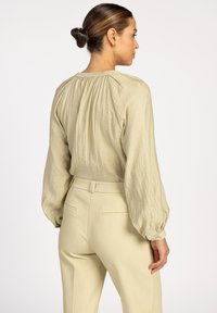 Helle beige Bluse mit Textur und weit ausgestellten Ärmeln, ausgestattet mit einem gerafften Rückenausschnitt. Kombiniert mit passenden, maßgeschneiderten beigen Hosen.