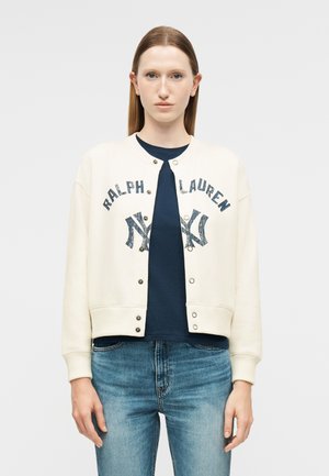 Mujer de cabello liso con una chaqueta blanca de botones Ralph Lauren sobre una camisa azul marino y jeans azules, mirando hacia adelante contra un fondo simple.