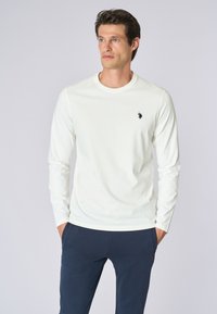 Maglietta bianca a maniche lunghe con scollatura rotonda e un piccolo logo nero sul petto, abbinata a pantaloni blu navy. Materiale in cotone liscio.