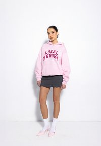 Pink hoodie with "LOCAL HEROES" text in dark pink, paired with a pleated black skirt. Worn with white socks and pink heels.