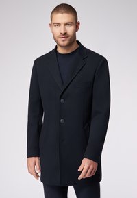 Cappotto di lana nera con colletto a punta, tre bottoni e taschino sul petto. Presenta una vestibilità sartoriale e una superficie testurizzata.