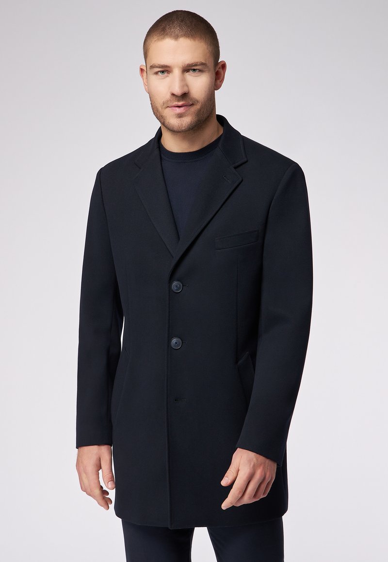 Cappotto di lana nera con colletto a punta, tre bottoni e taschino sul petto. Presenta una vestibilità sartoriale e una superficie testurizzata.