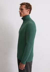 Pull zipé en maille côtelée verte avec col montant et manches longues, offrant une coupe décontractée. Associé à un pantalon gris pour un contraste.