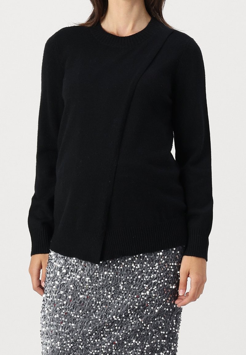 Kvinna som bär en svart långärmad tröja med diagonal sömdetalj och en kjol av silverpaljetter, stående mot en enfärgad bakgrund.