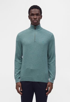 Homme portant un pull bleu sarcelle à fermeture éclair quart, manches longues, et un pantalon foncé, debout devant un fond blanc uni.
