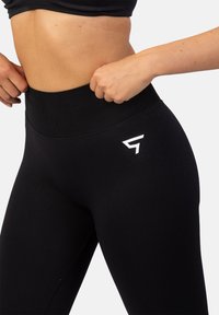 Čierne legíny s vysokým pásom a hladkým, elastickým povrchom. Obsahujú biele logo na boku a široký, pohodlný pás.