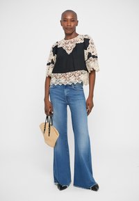 Top in pizzo nero e crema con maniche corte, abbinato a jeans svasati blu chiaro. Tiene una piccola borsa intrecciata, indossa scarpe nere.
