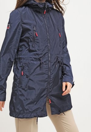 Kobieta ubrana w granatowy, długi, wodoodporny płaszcz z kapturem, zapinany z przodu na zamek, z licznymi kieszeniami na zamek oraz regulowanymi trokami w talii, w połączeniu z beżowymi spodniami.