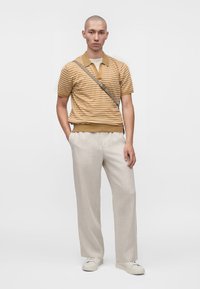 Homme portant un polo à manches courtes rayé moutarde et blanc, pantalon beige large, baskets blanches et sac bandoulière, debout avec une main dans la poche.