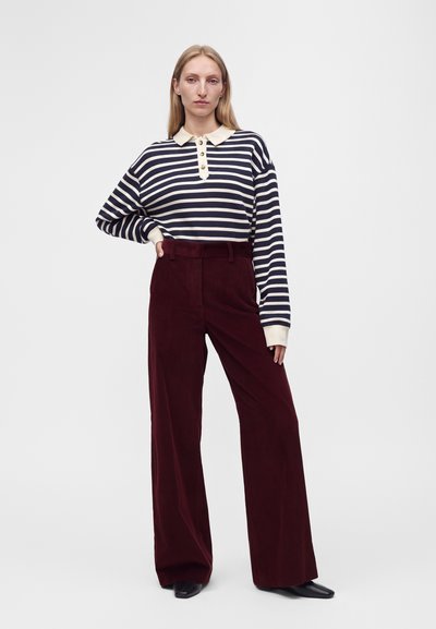 Femme portant un polo à manches longues rayé bleu marine et blanc et un pantalon large taille haute rouge foncé, debout devant un fond uni.